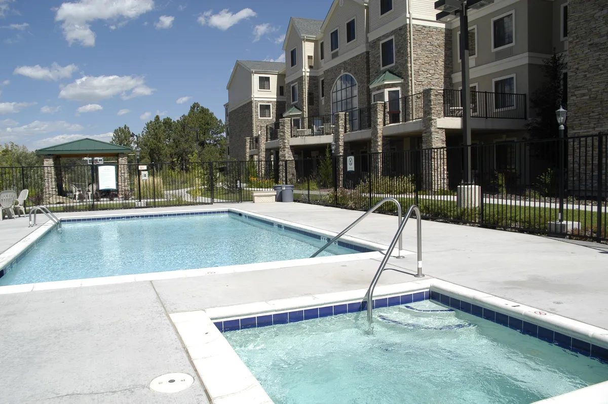 An inviting exterior pool area featuring a large swimming pool, adjacent hot tub, ample lounge chairs, and lush landscaping. The building facade is visible in the background, suggesting resort-style living. The scene exudes relaxation, with spaces for swimming, sunbathing, and socializing under the open sky.