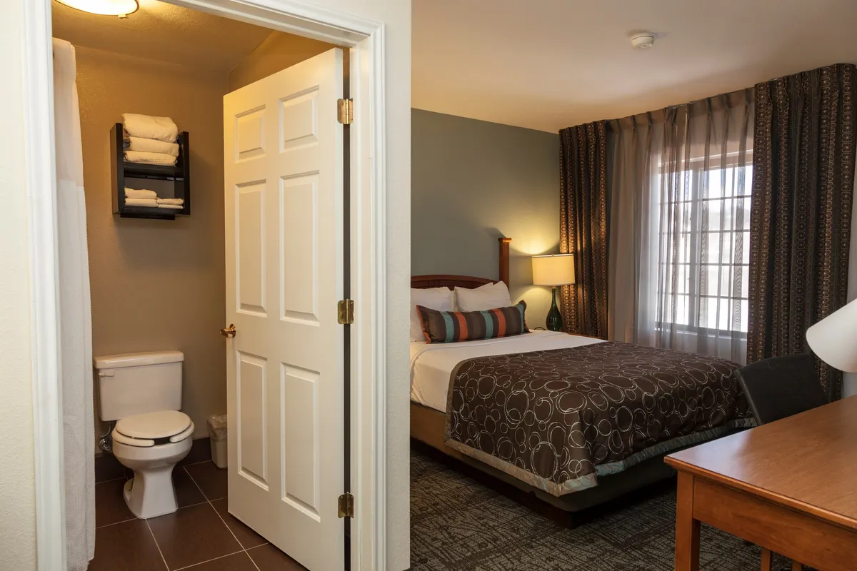 A cozy bedroom scene featuring a plush queen bed positioned to catch gentle natural light from the window. The room includes a desk ideal for reading or working, and the entrance to the bathroom is visible, subtly connecting the private sleeping area to the refreshing amenities beyond. Soft curtains and calm, comfortable decor evoke a welcoming, restful mood.